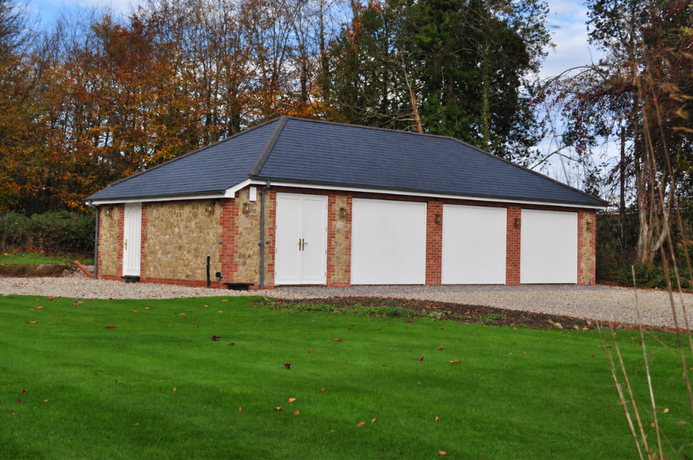 Bespoke built garage with three white garage doors, plus double and single doors, black tiled roof and bespoke brickwork.