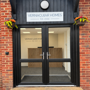 New black framed glazed double doors with Vernacular Homes sign above and two hanging baskets, with view to reception desk and signage on wall.